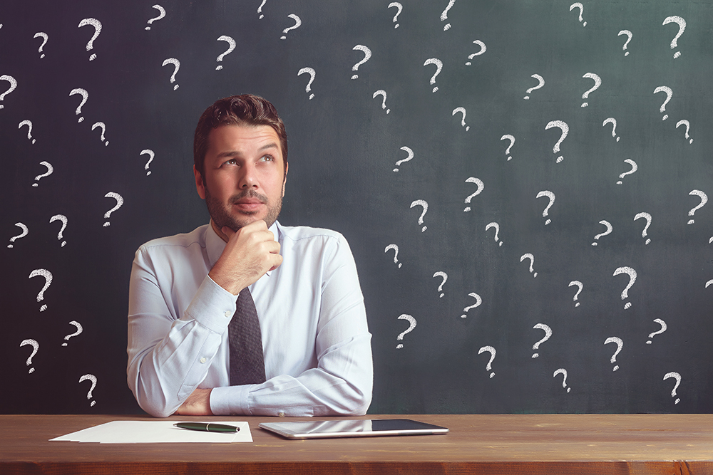 Man sitting at a desk with his hand on his chin thinking with question marks on the board behind him.