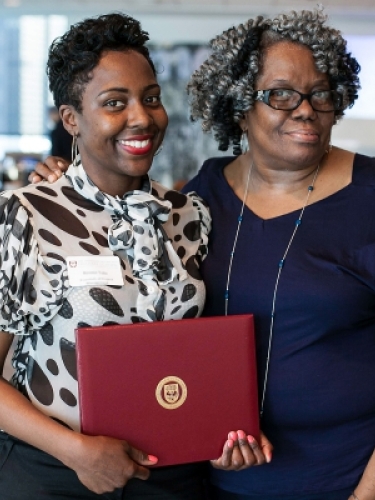Two people smiling at the camera during commencement. One student is holding a certificate.